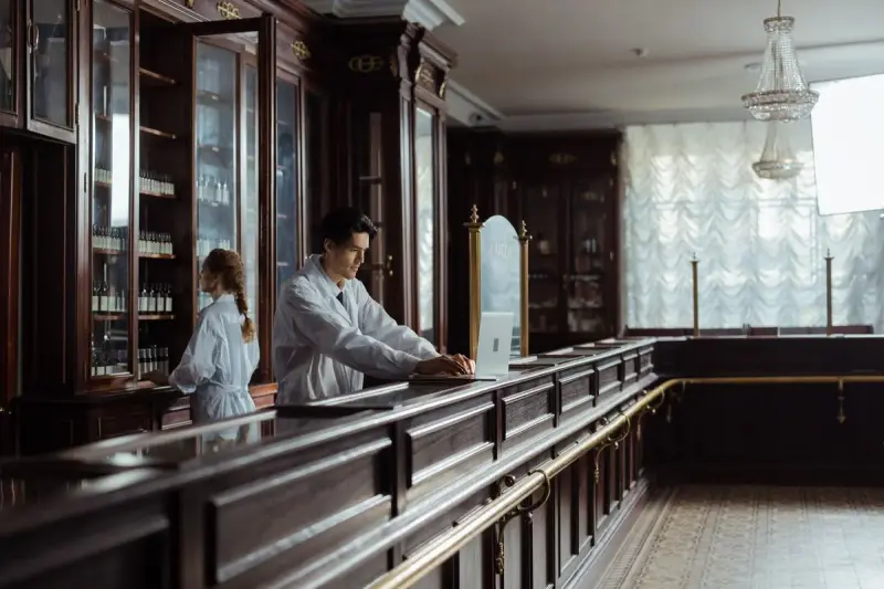 Long wooden pharmacy counter with glass cabinets, a pharmacist at a laptop and a colleague at the shelves