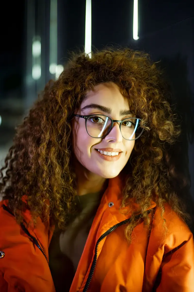 Smiling woman with curly hair in thin metal eyeglasses and an orange jacket at night