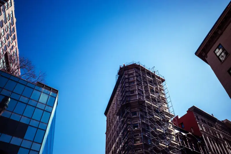 Brick high-rise wrapped in scaffolding next to a glass tower under a deep blue sky