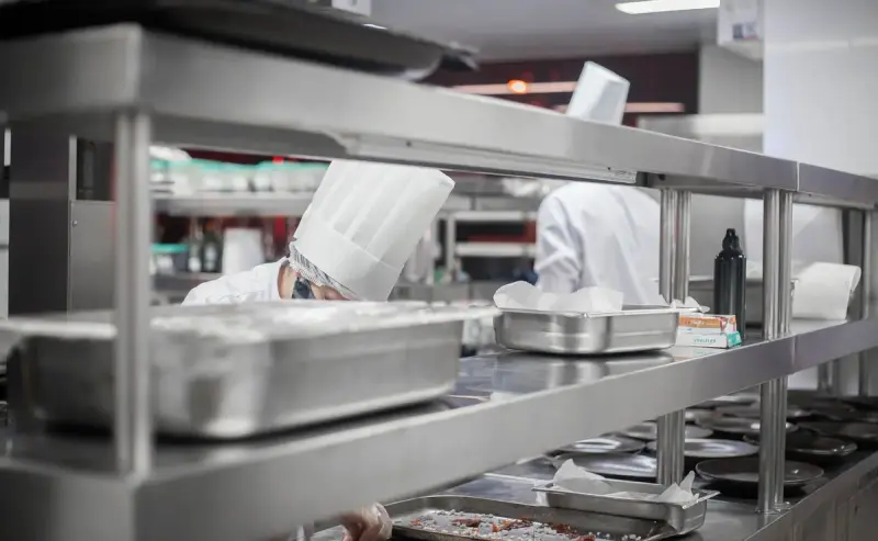 Chefs in white toques working beneath stainless steel shelving at a commercial kitchen pass