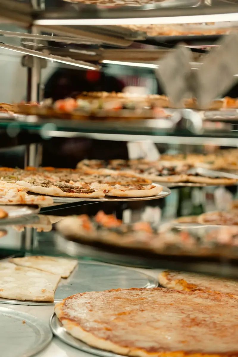 Whole New York style pizzas on metal trays inside a lit glass slice display case