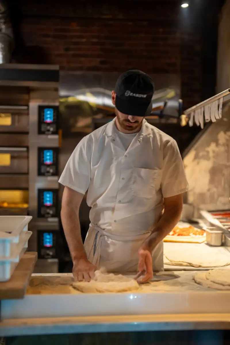 Pizza maker stretching dough on a marble counter in front of stacked electric deck ovens