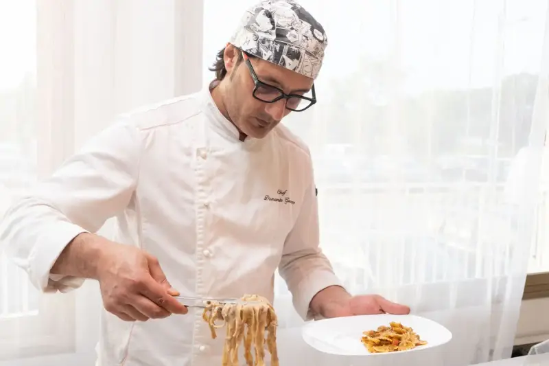 Chef in whites using tongs to plate fresh pasta in a bright dining room