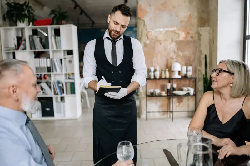 Waiter in a vest and tie taking an order from two diners at a restaurant table