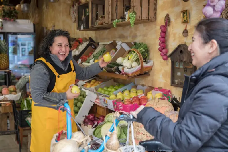 Laughing greengrocer in a yellow apron hands an apple to a customer beside crates of fresh produce