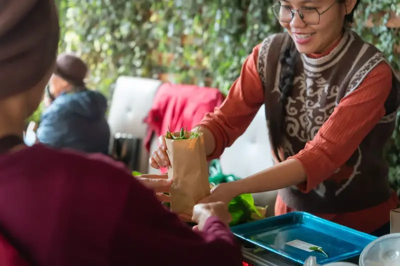 Smiling market vendor in glasses hands a paper bag of fresh greens to a customer at her stall
