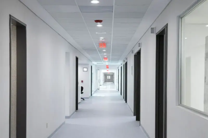 Long white hospital corridor with ceiling lights, exit signs and glazed doors