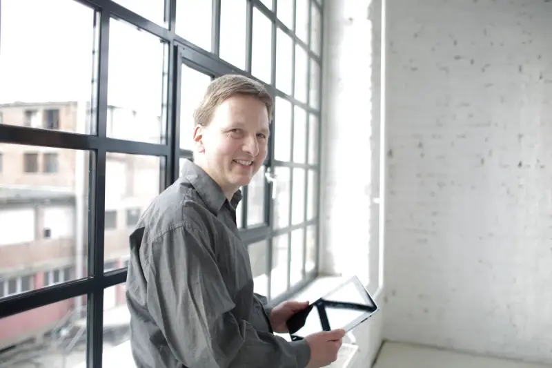 Smiling manager holding a tablet beside tall steel-framed windows in a bright workspace