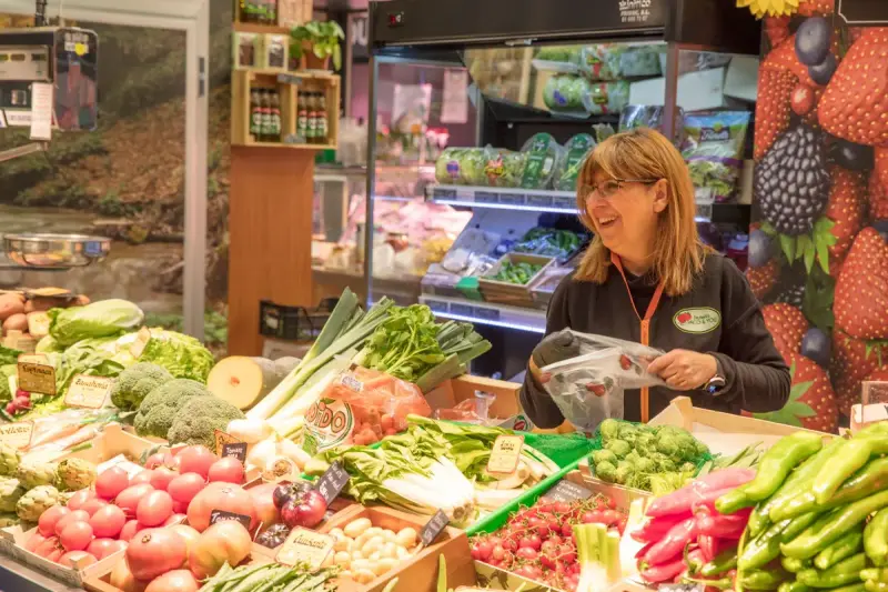 Smiling produce clerk bags vegetables behind a full counter of tomatoes, peppers, leeks and broccoli