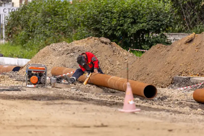 Worker welding a long steel pipe beside a trench on a sunny site
