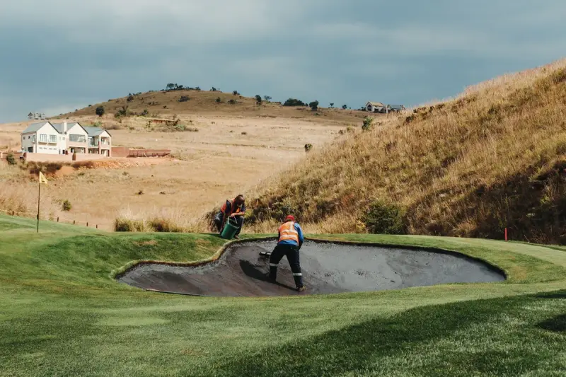 Grounds crew raking a sand bunker on a golf course below rolling hills