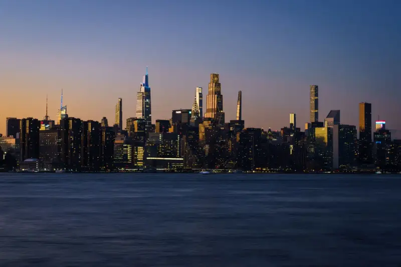 Manhattan skyline at dusk across the water with towers lit against a deep blue sky
