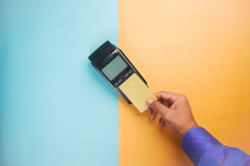 Hand tapping a card on a black payment terminal against a blue and orange backdrop