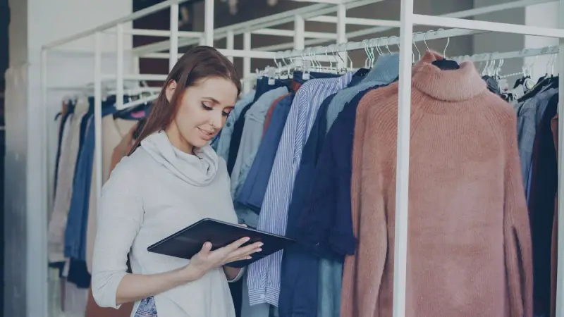 Shop assistant checking inventory on a tablet beside racks of sweaters and coats