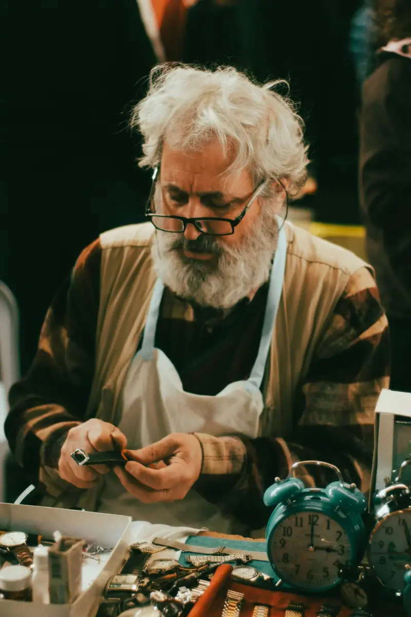 Senior watchmaker in an apron repairing a timepiece among vintage alarm clocks
