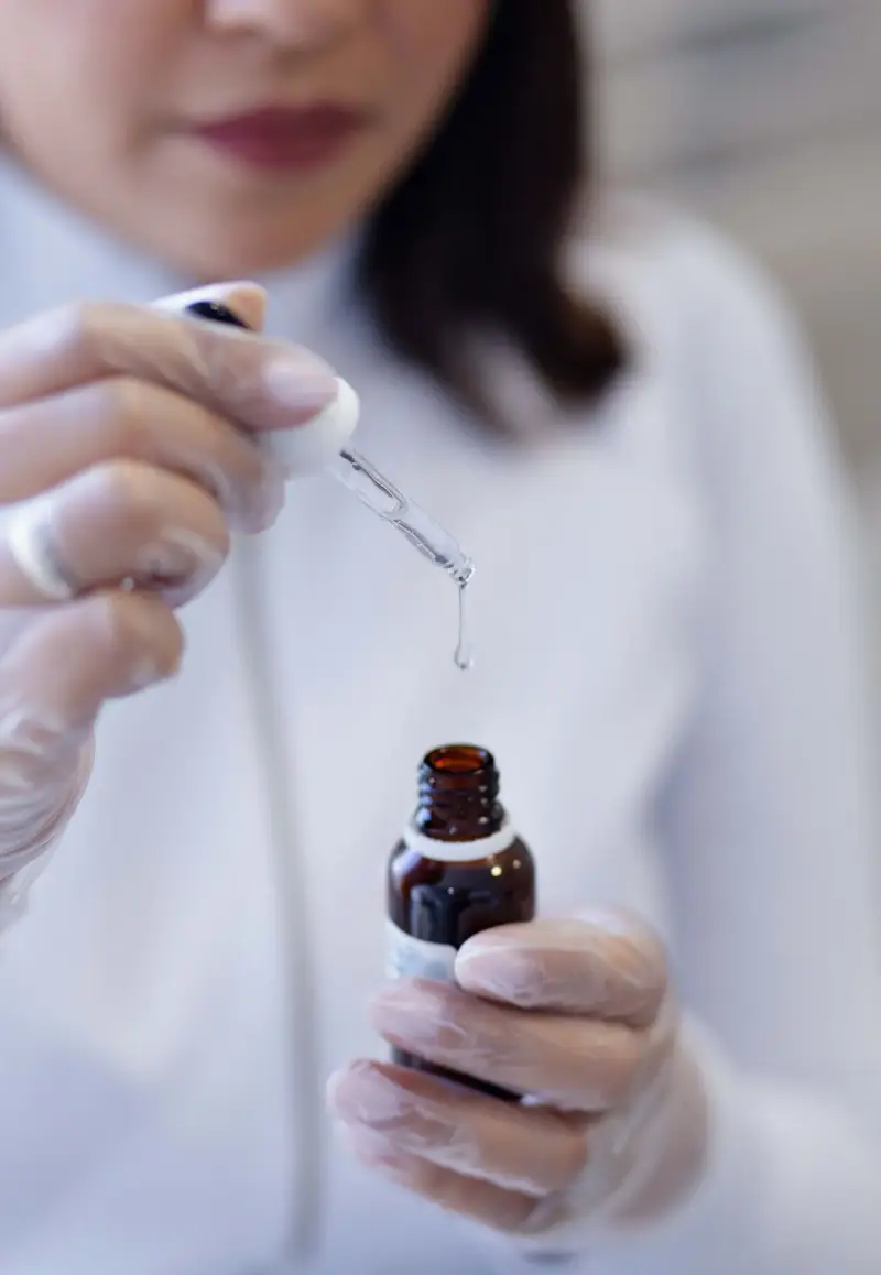 Gloved hands dropping serum from a pipette into an amber glass bottle