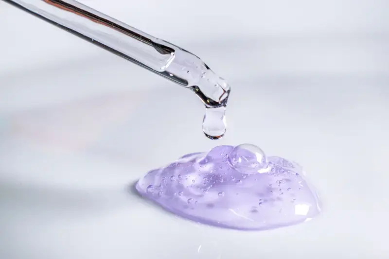 A pipette releases a drop onto a swatch of lilac skincare gel on a white surface