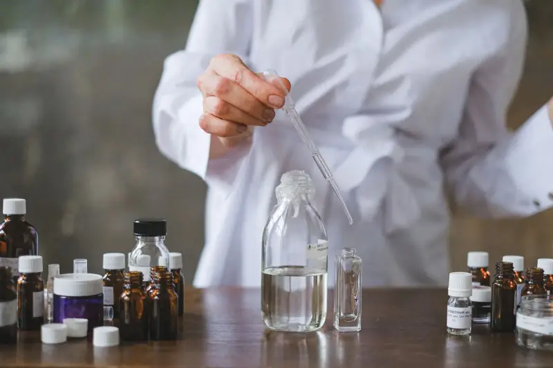 Chemist in a white coat using a dropper over glass bottles on a wooden lab table