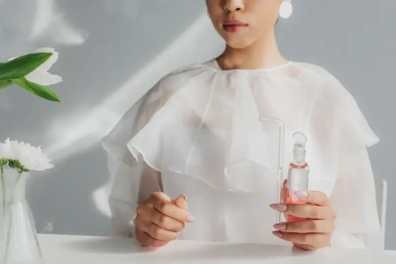 Woman in a white blouse holding a small glass flask of pink liquid at a minimalist desk