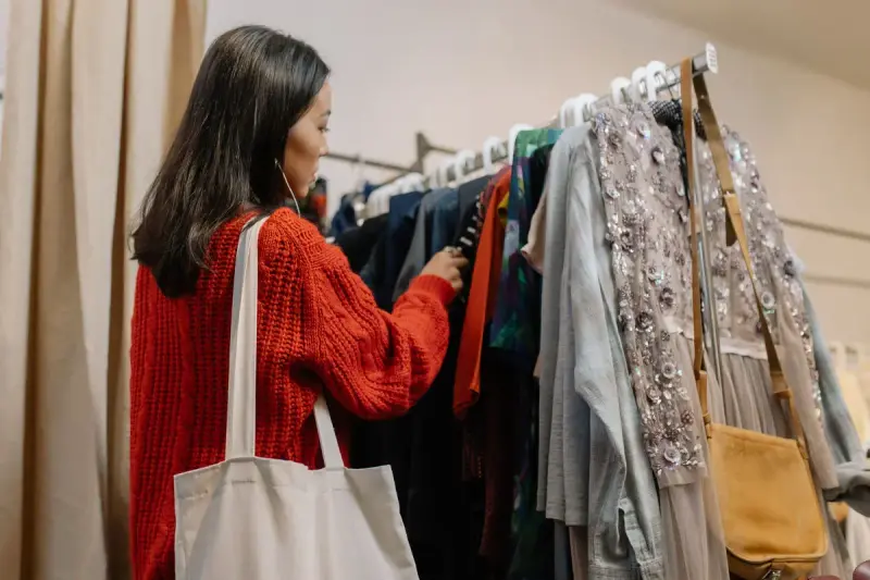Woman in a red knit sweater with a canvas tote flipping through a packed clothing rack
