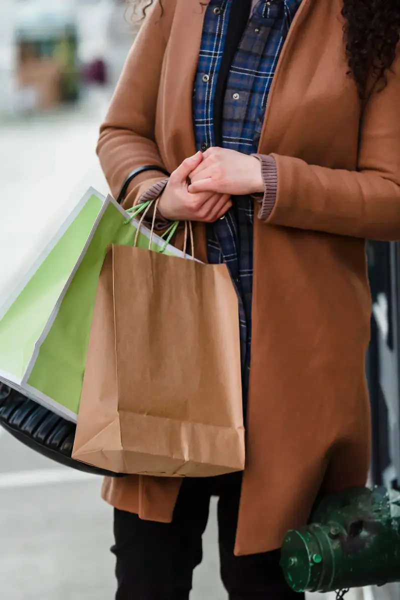 Woman in a camel coat and plaid shirt carrying paper shopping bags on a city street