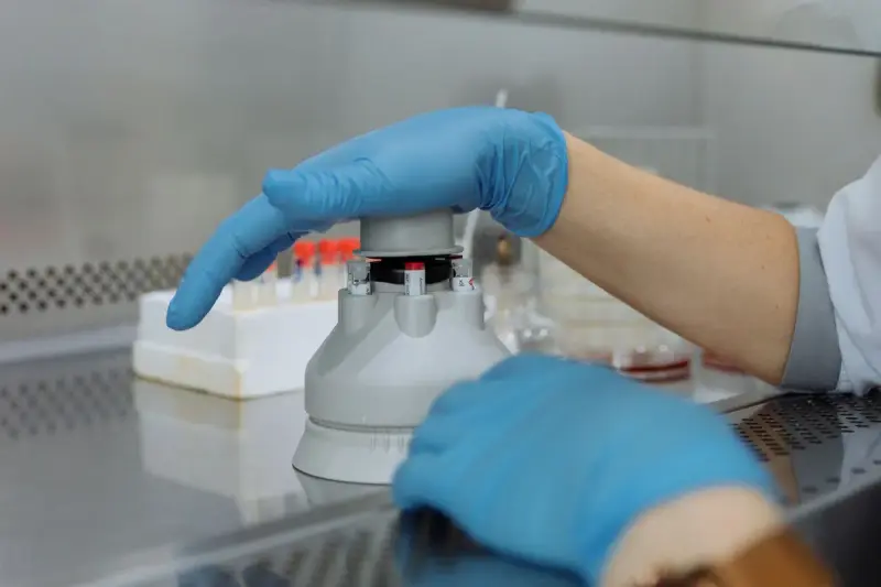 Gloved hands loading tubes into a benchtop centrifuge with lab glassware in the foreground