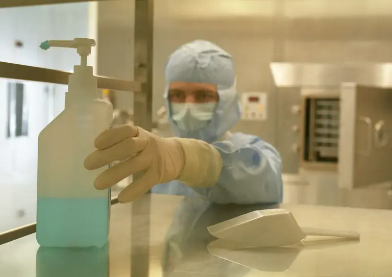 Operator in a full cleanroom gown, mask and gloves reaching for a dispenser bottle