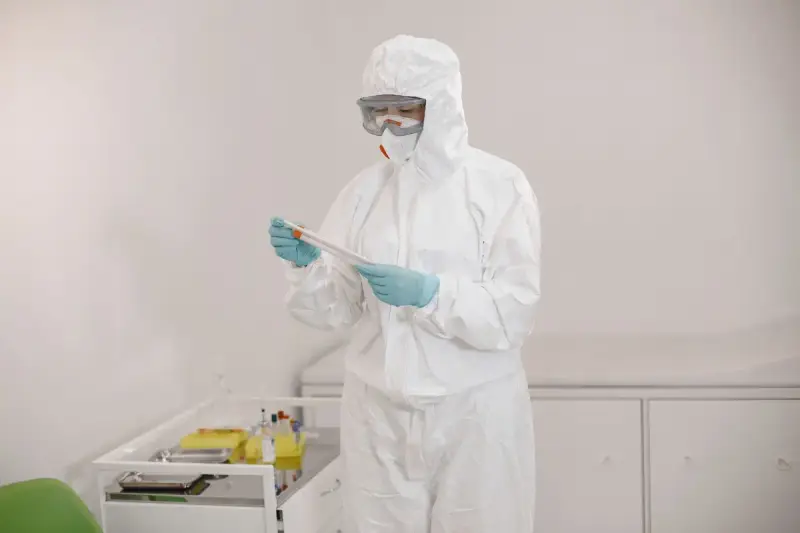Scientist in a white coverall, goggles and gloves examining a sample tube in a lab