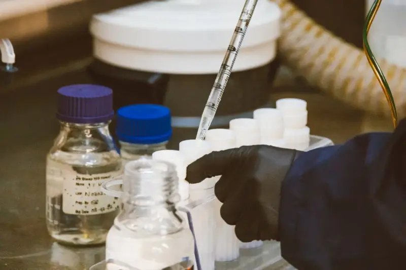 Gloved hand using a pipette among reagent bottles and sample tubes on a lab bench