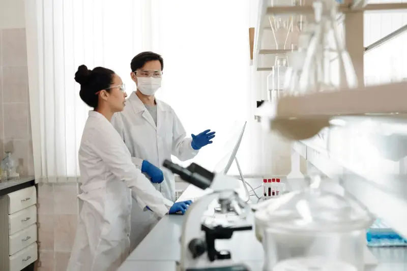 Two scientists in lab coats and masks discussing work at a bench with a microscope