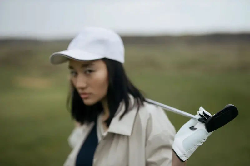 Woman golfer in a white cap and beige jacket holding a club on a course