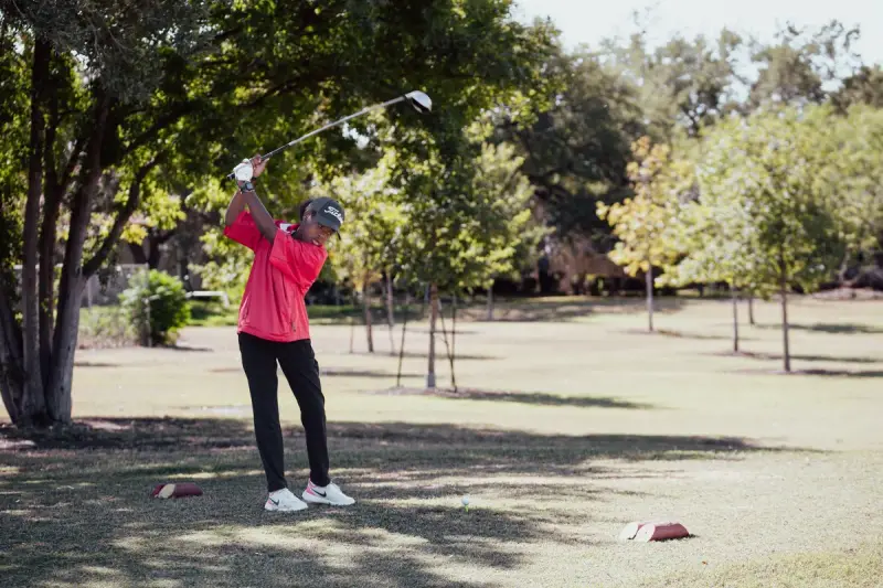 Young golfer in a coral polo swinging a driver under shade trees
