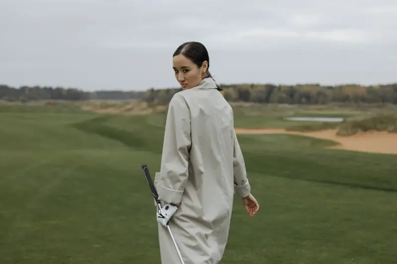 Woman in a long coat walking across a links-style course holding a club