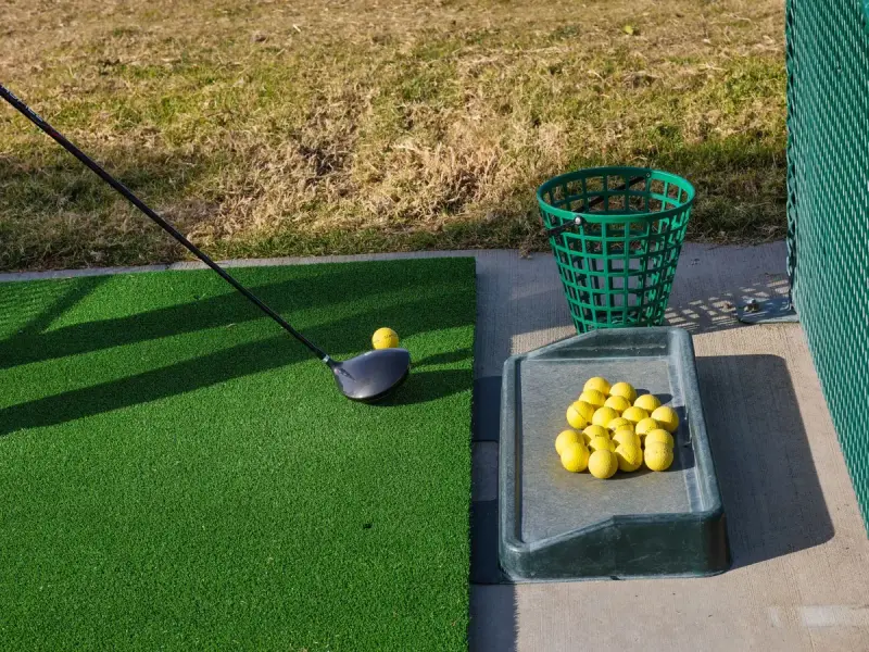 Driver resting behind a yellow range ball on a green hitting mat beside a ball tray