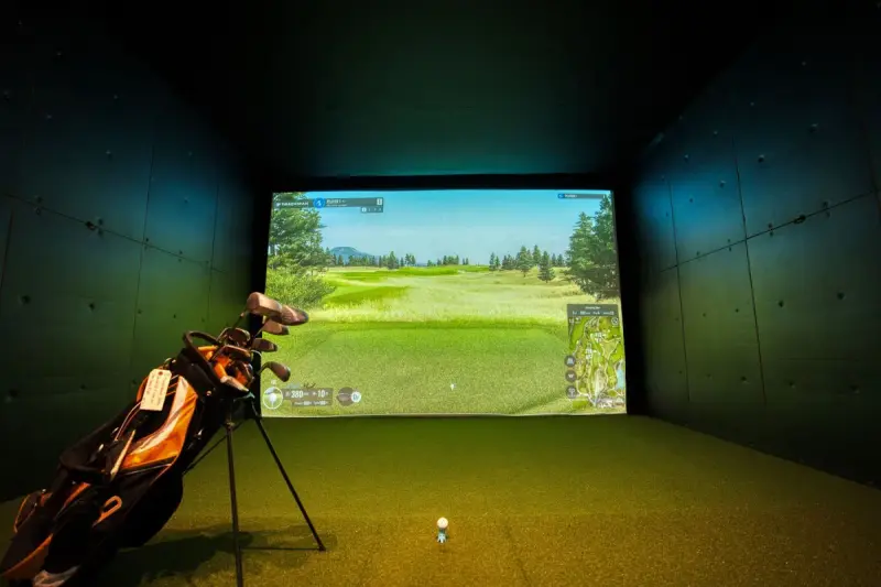 Indoor golf simulator bay with a projected fairway on screen and a bag of clubs on turf