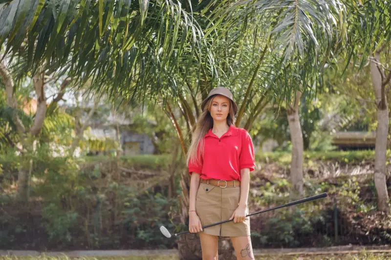 Woman golfer in a red polo and bucket hat holding a club under palm trees