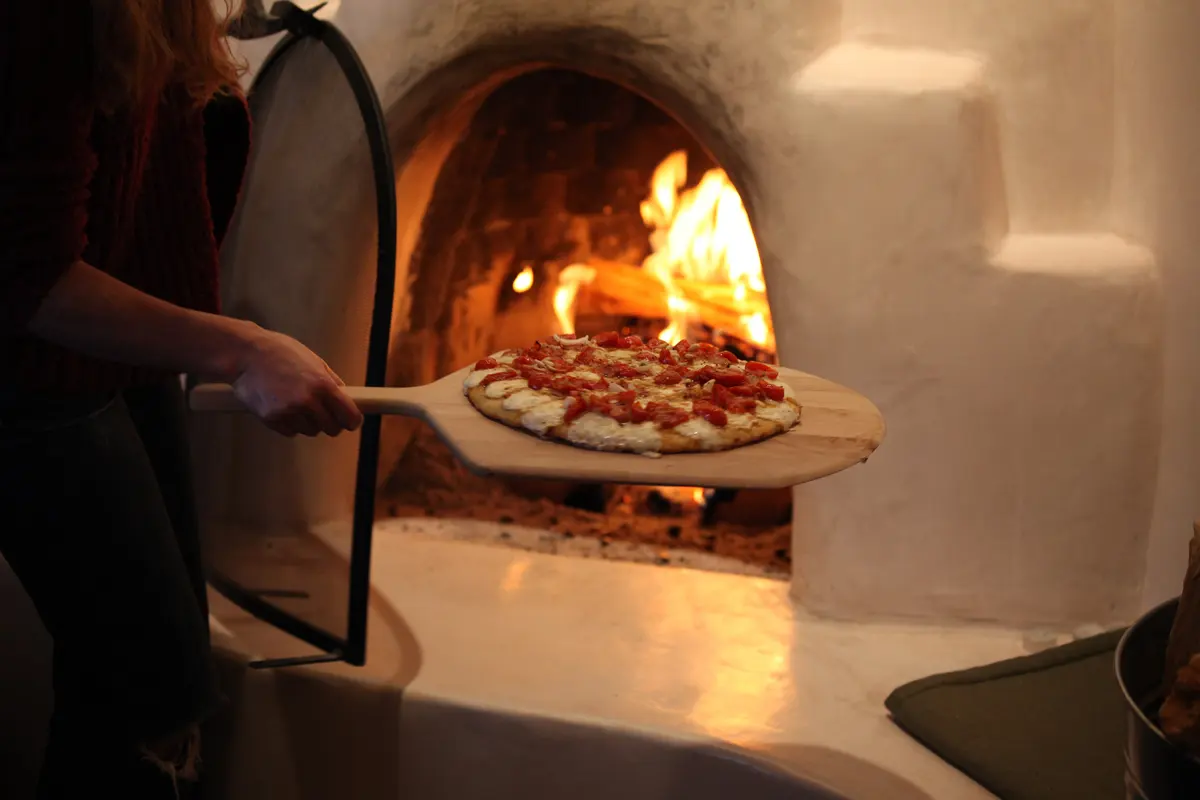 Pizza with tomatoes on a wooden peel going into a glowing wood-fired oven