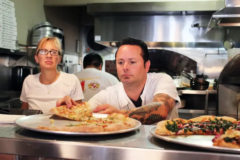 Pizza instructor tasting a student's test pizza at a stainless kitchen counter