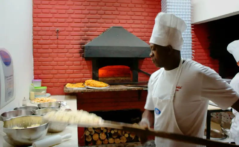 Pizzaiolo in a white hat working a peel in front of a wood-fired oven