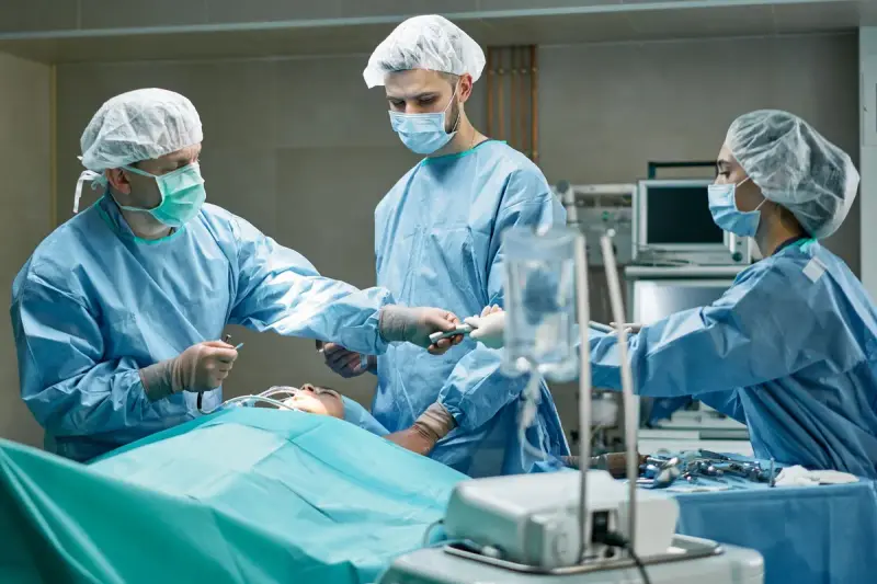 Three surgeons in blue gowns and caps working over a draped patient in an operating room