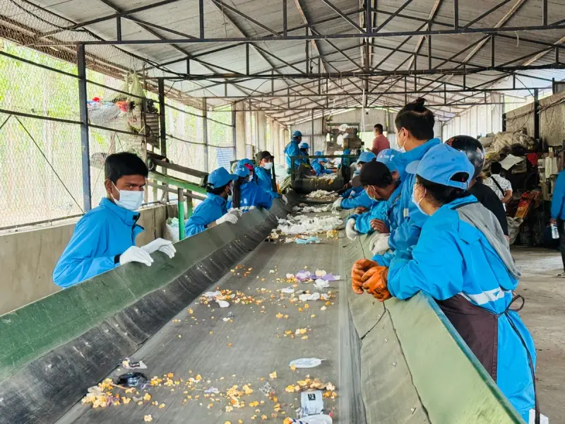 Workers in blue uniforms and caps sorting recyclables along a long conveyor under a steel roof