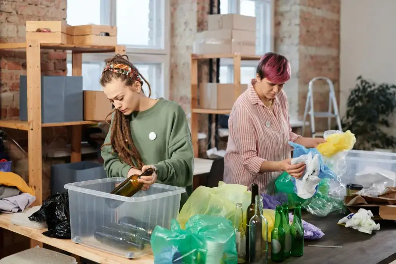 Two women sorting glass bottles and plastic bags into bins at a workshop table