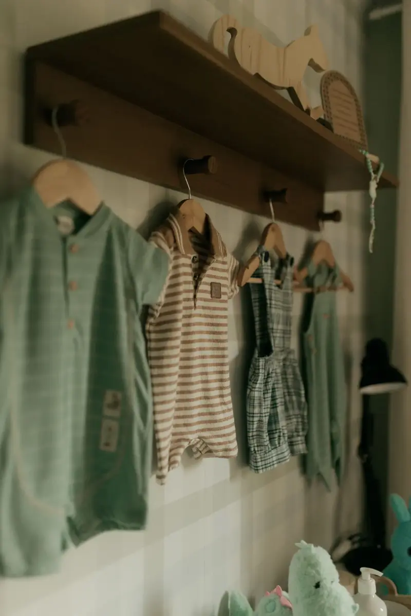 Sage, striped and plaid baby rompers hanging on wall pegs under a wooden shelf