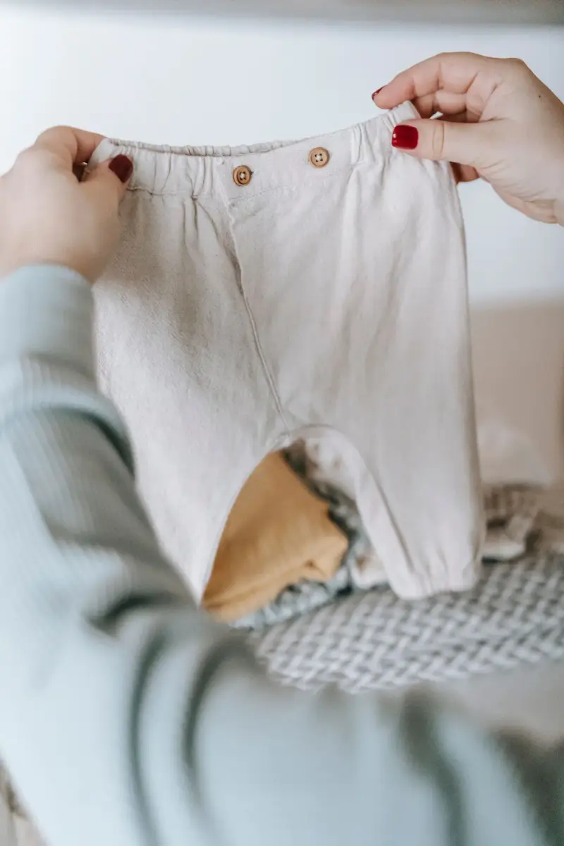 Hands holding small linen baby trousers over folded garments