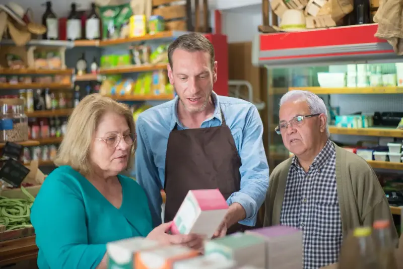 Grocery clerk in a brown apron shows a boxed product to two older shoppers in a well-stocked store