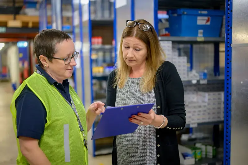 Two women review an inventory checklist on a clipboard between warehouse shelves of boxed stock
