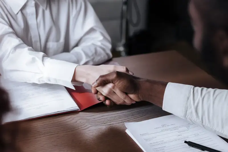 Two people shaking hands over a desk with documents and a pen