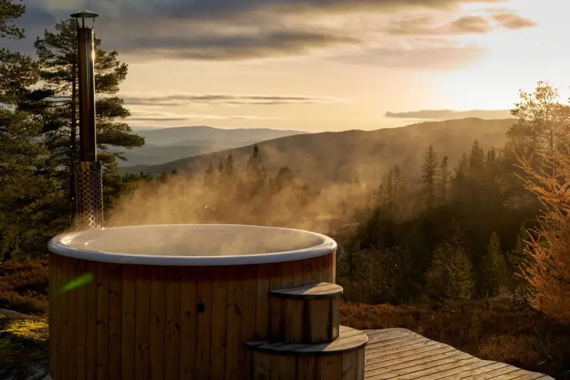 Steaming wood-fired hot tub on a deck above a forested valley at sunset