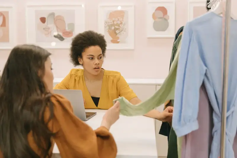 Women at a table reviewing a fabric swatch beside a rack of shirts