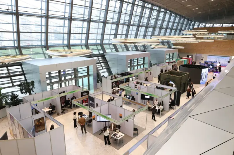 Exhibition hall seen from above with rows of white stands and visitors walking the aisles